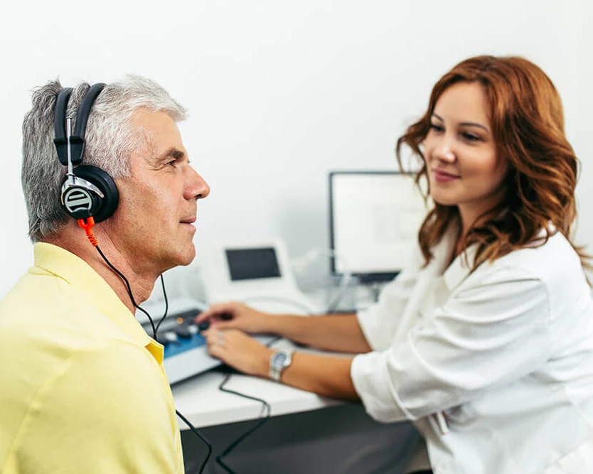 Profissional realizando um exame de audição em um paciente que usa fones, em frente ao equipamento